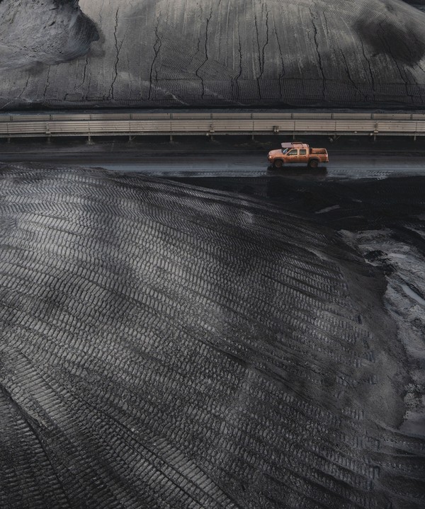 Kohlenlager mit Reifenspuren. Im Hintergrund ein oranges Baufahrzeug, das klein auf der großen schwarzen Fläche wirkt.