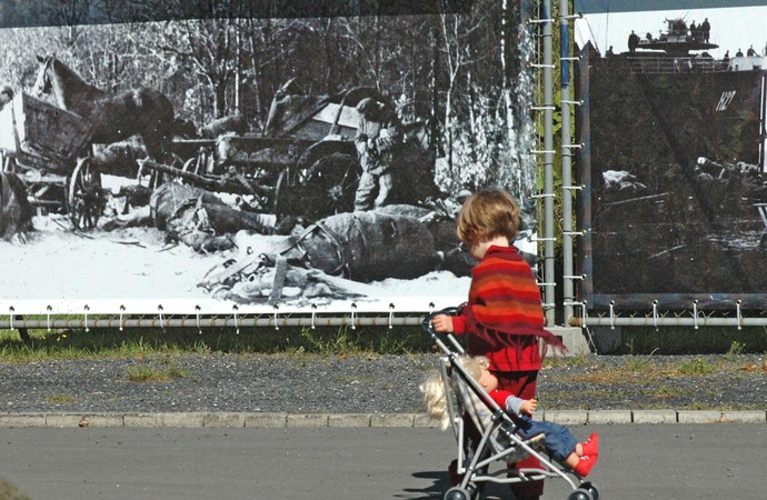 Mädchen mit Puppenwagen vor einem historischen Foto eines Flüchtlingstrecks.