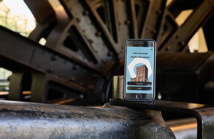 Smartphone mit geöffnetem Mediaguide steht vor dem Rad der Dampffördermaschine