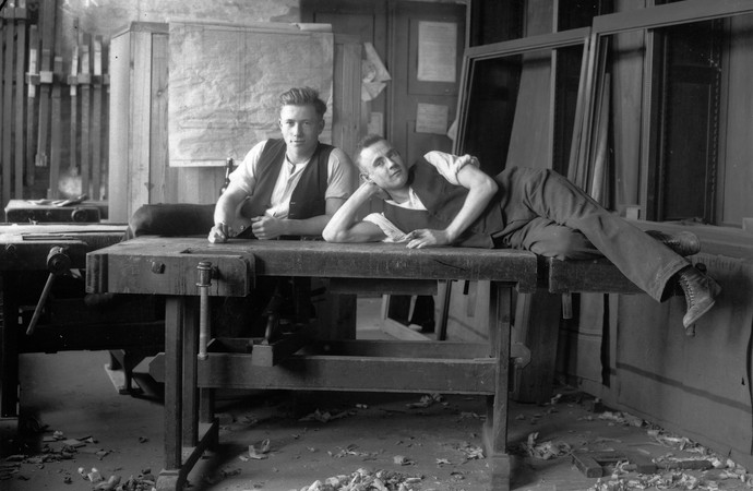 Zwei junge Schreinergesellen liegen auf einer Hobelbank. Historische Schwarz-Weiß-Aufnahme.