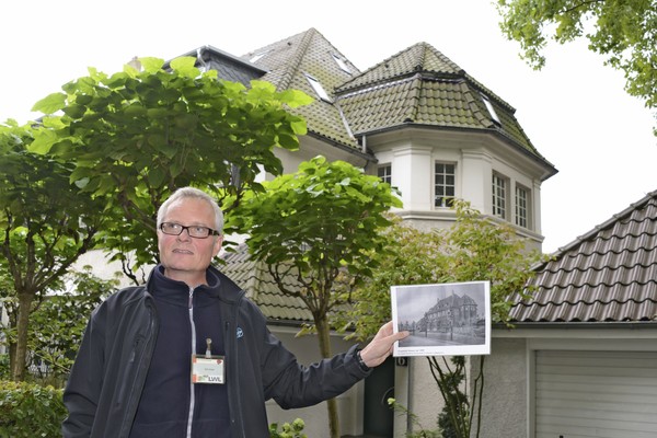 Ein Gästeführer steht vor einem Siedlungshaus und hält ein historisches Foto in die Höhe.
