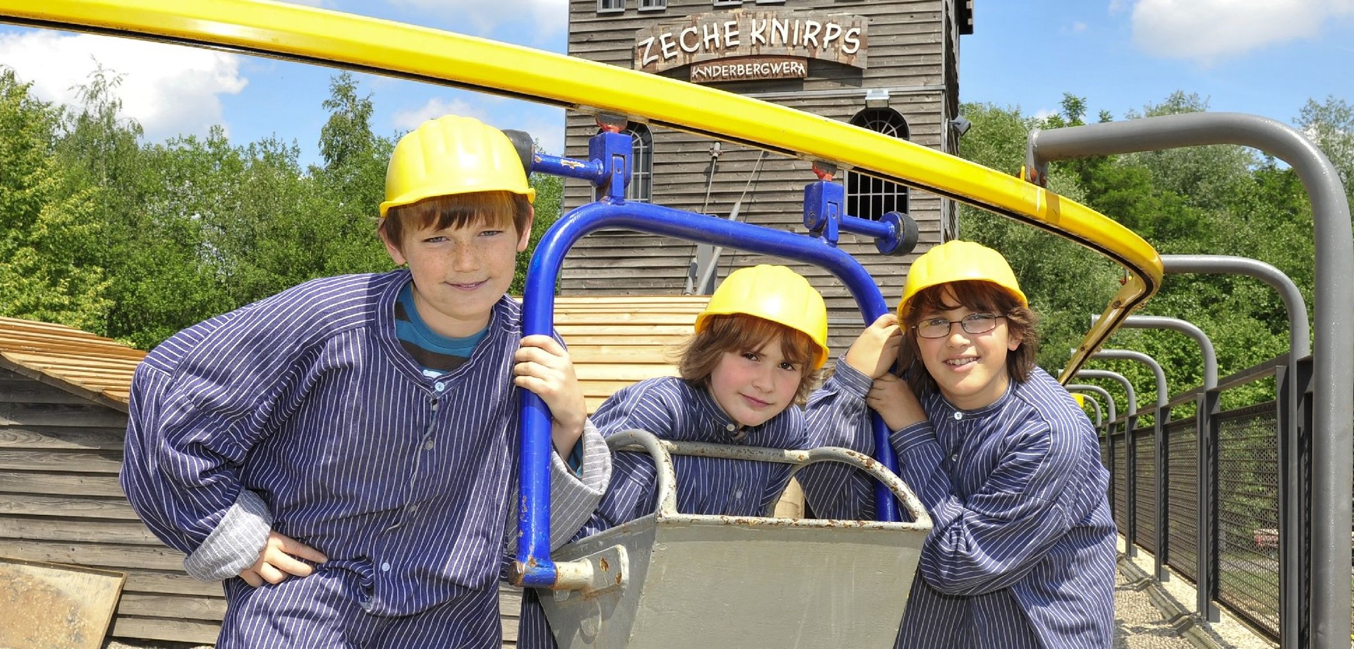 Drei Kinder in Bergarbeiter-Kleidung an einer Lore auf dem Bergbauspielplatz "Zeche Knirps"