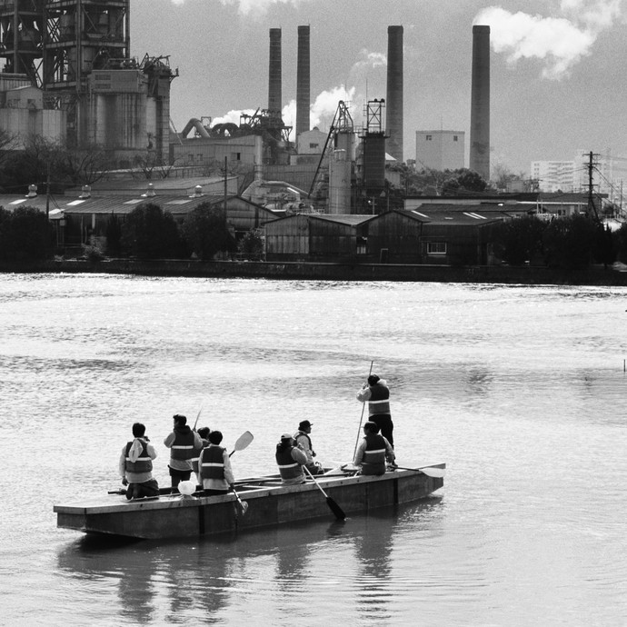 Ein Boot mit 8 Personen auf einem Fluss, Im Hintergrund eine Industrielandschaft mit verschiedenen Gebäuden und rauchenden Schornsteinen. (vergrößerte Bildansicht wird geöffnet)