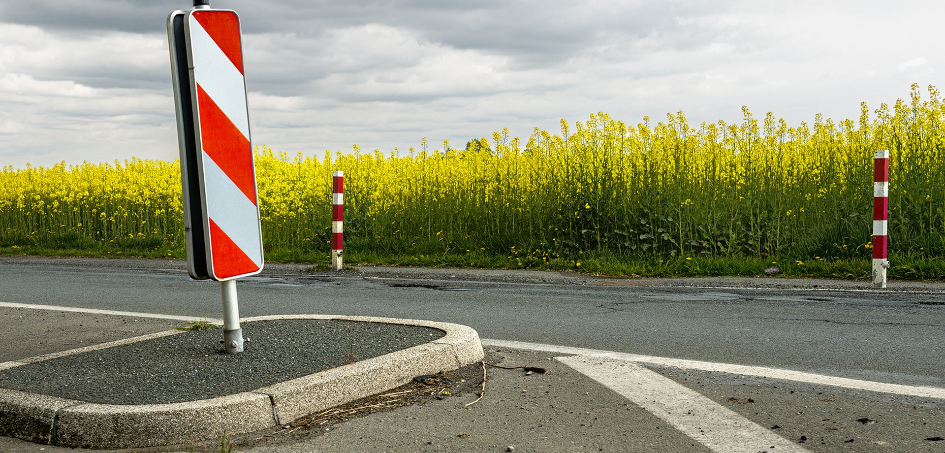 Aufnahme einer Straße mit einer kleinen Verkehrsinsel, auf der eine rot-weiße Warnbarke steht. Im Hintergrunde in Rapsfeld.